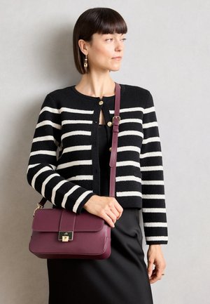 Kvinde iført sort og hvid stribet cardigan og sort nederdel, der holder en lilla læder crossbody taske med guldlås.