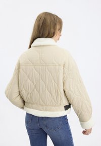 Veste matelassée beige avec un col et des poignets en fourrure blanche, présentant un design ample et un motif texturé, assortie à un jean bleu.