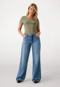 Olive green fitted t-shirt with "Guess est.1981" print, paired with light blue wide-leg denim jeans. Smooth texture, casual design.