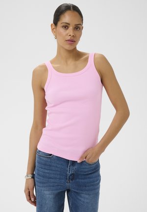 Frau trägt ein ärmelloses hellrosa Oberteil und blaue Jeans, mit der rechten Hand in der Tasche, vor einem einfarbigen grauen Hintergrund.