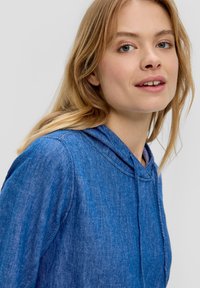 Blauwe hoodie van een zachte, textuurachtige stof. Voorzien van een trekkoordkap en een ontspannen pasvorm, benadrukt door subtiele stikdetails.