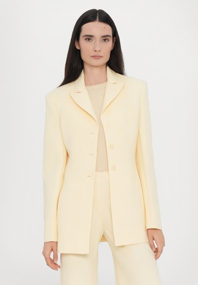 Light yellow tailored blazer with a notch lapel, single-breasted design, and five buttons, paired with a beige top and matching trousers.