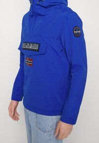 Muž v jasně modré bundičce Napapijri s přední kapsou a nášivkami, doplněné světle modrými džíny.