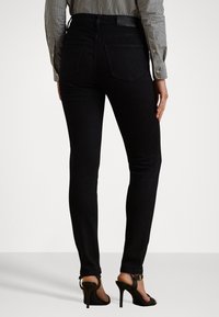 Jeans skinny in denim nero con vita alta e tasche posteriori. Il modello indossa sandali neri con tacchi alti e una maglietta a righe.