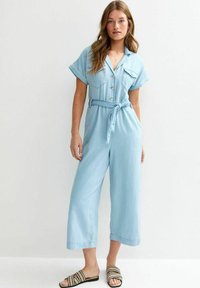 Combinaison en denim bleu clair avec des manches courtes, fermeture par boutons, deux poches poitrine et une ceinture à nouer à la taille. Design à jambes larges, texture douce.