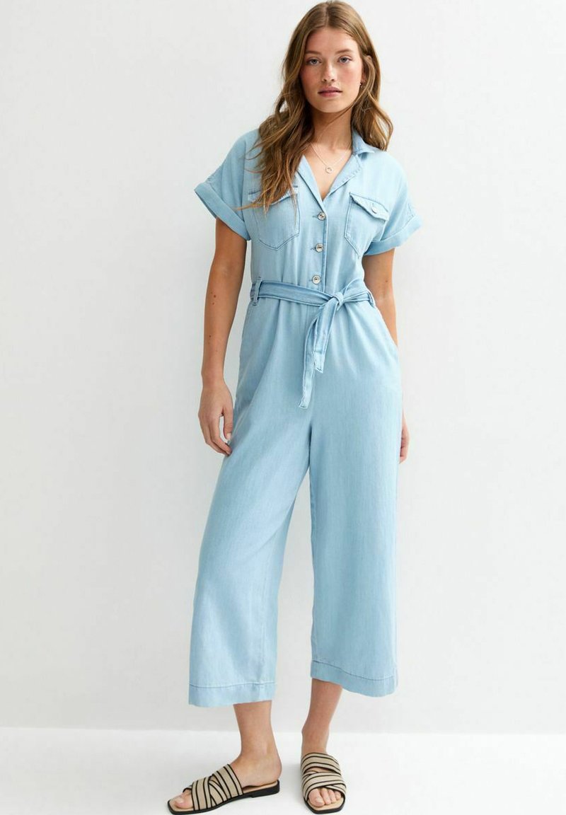 Combinaison en denim bleu clair avec des manches courtes, fermeture par boutons, deux poches poitrine et une ceinture à nouer à la taille. Design à jambes larges, texture douce.