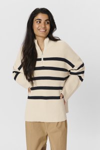 Kreemja ja tumesinise triibuga pullover-sweater poole tõmbe lukuga krae, ribakudumise tekstuuriga ja lahtise sobivusega, koos beežide pükstega.