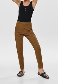 Brown tapered pants made of stretch fabric with front pockets, paired with a black tank top and black slide sandals featuring studded accents.