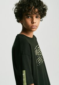 Czarna bawełniana koszulka z grafiką, na której widnieją poziome paski oraz naszywka na lewym rękawie z zielonymi akcentami.
