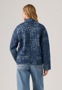 Quilted blauwe denimjas met paisleypatroon, geribbeld textuur en een hoge kraag. Achteraanzicht toont contrasterende stiksels en een ontspannen pasvorm.