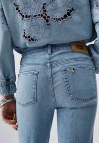 Gros plan sur l'arrière d'un jean en denim bleu clair avec deux poches et un patch de marque en cuir marron, accompagné d'un haut bleu orné de broderies florales découpées.