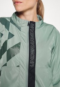 Ljusgrön vindjacka med en svart vertikal zip och logotyp. Har geometriska mönster i mörkare grönt och två sidfickor.
