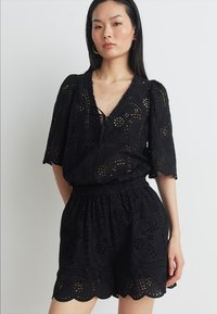 Romper noir en dentelle à œillets, présentant un décolleté en V, des manches courtes bouffantes, une taille élastique et un ourlet festonné, avec des motifs floraux intriqués.