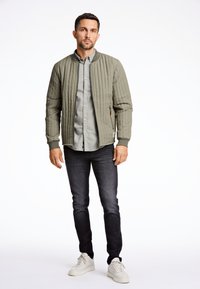 Chaqueta bomber acolchada de color verde oliva con puños de canalé, camisa de botones gris, jeans oscuros y zapatillas blancas. Textura suave, diseño minimalista.