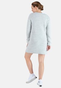 Robe tricotée gris clair à manches longues, avec des poignets côtelés, un ourlet arrondi et un petit logo près de l'encolure arrière. Portée avec des baskets blanches.