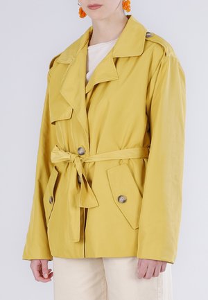 Persona che indossa una giacca color senape con cintura, tasche con patta e bottoni, e orecchini arancioni, in piedi davanti a uno sfondo semplice.