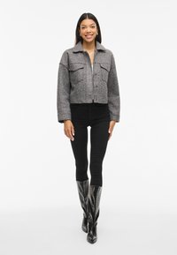 Grå jakke i kort modell med to brystlommer, krage og strukturert stoff, parret med svarte leggings og knelange svarte støvler.