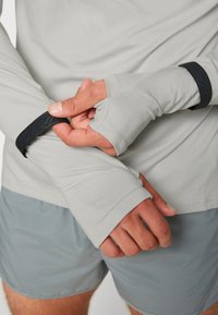 Camiseta de manga larga gris claro con agujeros para los pulgares y tiras negras como acento. Presenta una tela suave y un diseño ajustado, se muestra junto con pantalones cortos grises.