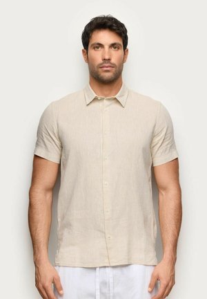 Mężczyzna stojący przodem ubrany w beżową, krótko rękawową koszulę na guziki i białe spodnie na jednolitym jasnym tle.