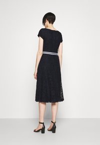 Žena s krátkými tmavými vlasy oblečená v černých krajkových midi šatech s bílým páskem, černých sandálech na blokovém podpatku s kotníkovým řemínkem, stojící proti jednoduché zdi.