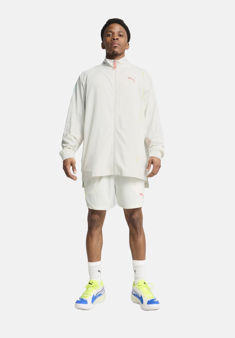 Veste légère zippée beige pâle avec des accents roses, associée à un short assorti. Les chaussures présentent des détails bleu, blanc et vert fluo.