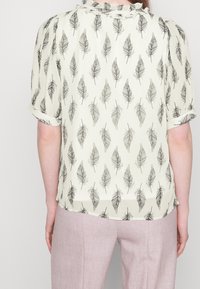 Blouse crème avec motif de feuilles noires, col froncé et manches courtes. Portée avec un pantalon rose clair. Texture lisse.