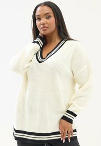 Sweter z dzianiny w kolorze kremowym, z dekoltem w kształcie V oraz czarnymi paskami na kołnierzu i mankietach, z długimi rękawami i luźnym fasonem.