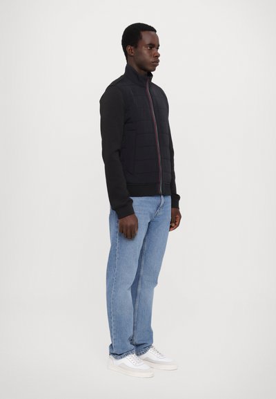 Svart jakke med quiltet front, høy krage og glidelås; kombinert med blå jeans og hvite sneakers, på en nøytral bakgrunn.