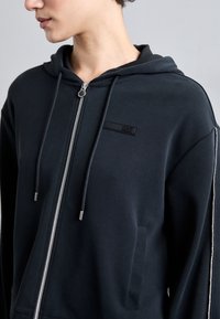 Felpa con zip di colore blu scuro realizzata in tessuto morbido, con un piccolo logo nero, cerniera argento, cordini e tasche laterali.