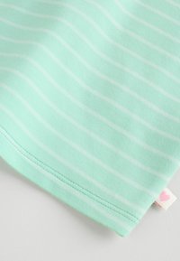 Lysegrønt stof med tynde hvide vandrette striber og detaljerede sømme, der har en lille mærkat med et pink hjerte symbol.