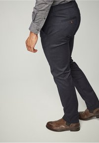 Man som bär mörkblå kostymbyxor, bruna läderskor och en grå långärmad skjorta med knappar, står mot en enkel bakgrund.