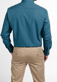 Turquoise geruit overhemd met lange mouwen en knoopmanchetten, gecombineerd met lichtbeige broeken met achterzakken met knopen.