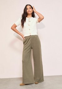 Balta īsajām piedurknēm kardigāns ar zeltīšiem pogām, saderēts ar vaļīgām olīvzaļām ribota auduma biksēm. Mīksta tekstūra un brīvs dizains.