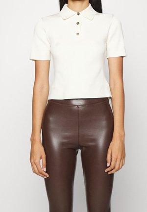 Personne portant un polo blanc côtelé à manches courtes avec trois boutons et un pantalon en cuir marron taille haute, debout devant un fond uni.