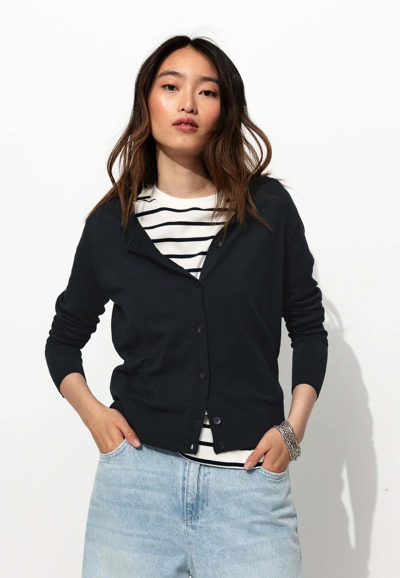 Cardigan noir avec boutons sur le devant et manches longues, porté sur une chemise rayée. Jean en denim bleu clair avec une coupe décontractée, les mains dans les poches.
