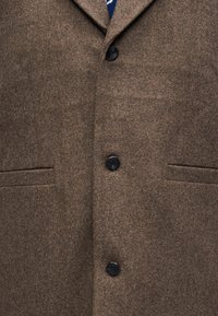 Καφέ μάλλινο παλτό με απαλή υφή, με γιακά με σχισμή, τρία μαύρα κουμπιά και δύο μπροστινές τσέπες.