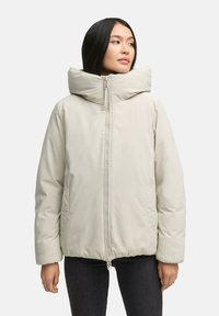 Donna che indossa un piumino beige chiaro con colletto alto e zip frontale, abbinato a pantaloni scuri, mentre guarda di lato.