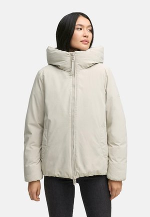 Vrouw in een lichtbeige pufferjack met een hoge kraag en een ritssluiting aan de voorkant, gecombineerd met donkere broek, kijkt opzij.