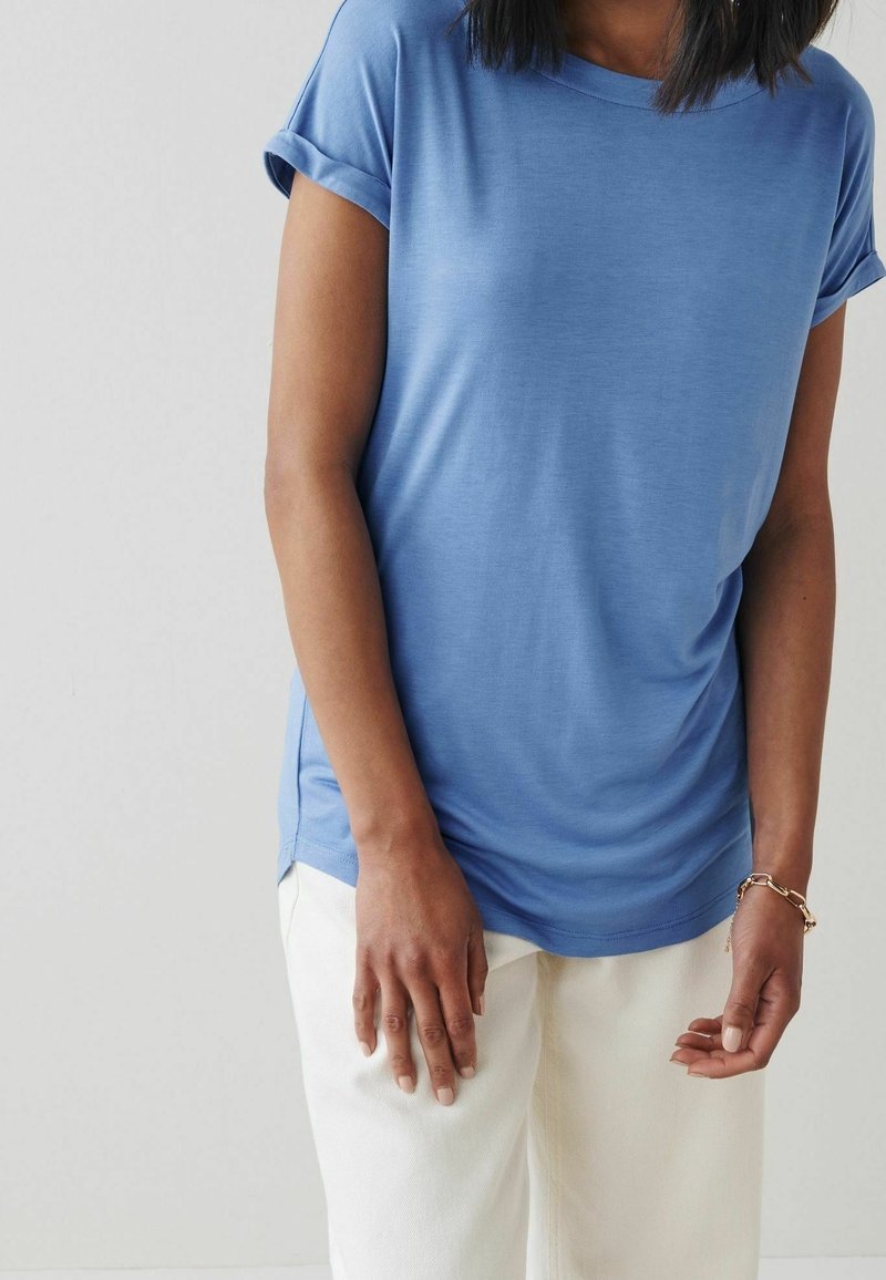 Femme portant une chemise bleue à manches courtes et un pantalon blanc, avec un bracelet chaîne en or à son poignet droit, debout devant un fond uni.