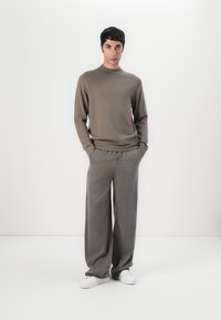Maglione di colore marrone a maglia con un collo alto, abbinato a pantaloni grigi a gamba larga e sneakers bianche, presentato su uno sfondo neutro.