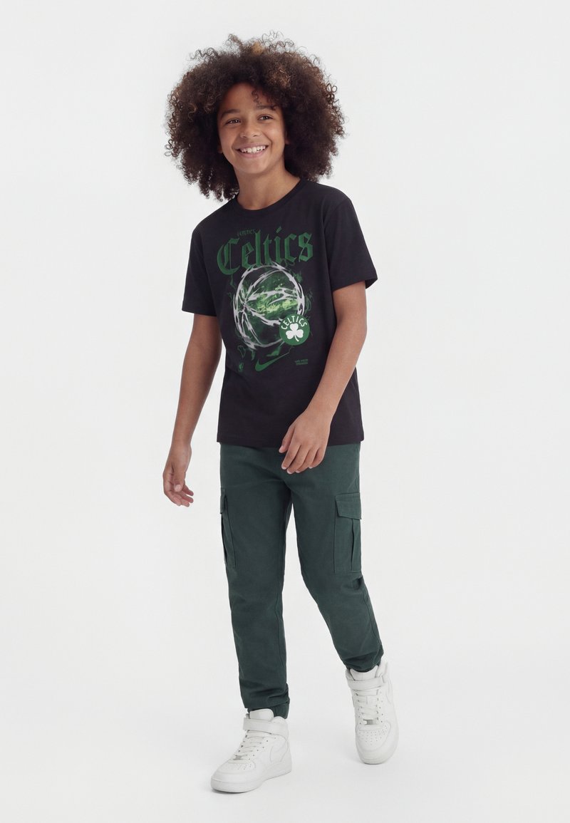 Jongen met krullend haar die glimlacht, lopend in donkergroene cargobroek, zwart T-shirt met Celtics-opdruk en witte sneakers tegen een effen achtergrond.