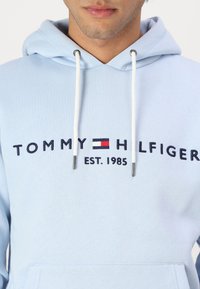 Detail osoby v světle modré mikině s bílými šňůrkami a výšivkou loga "Tommy Hilfiger EST. 1985" na hrudi.