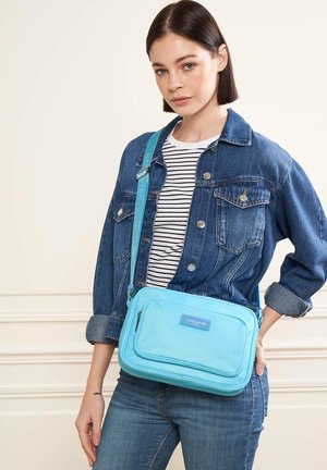 Sac en bandoulière bleu avec une texture lisse, des poches zippées et une sangle réglable, porté sur une veste en denim et une chemise rayée.
