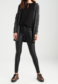 Svart blus med knytning vid halsen, grå lång kofta, byxor i läderliknande material med räfflade detaljer och bruna snörskor.