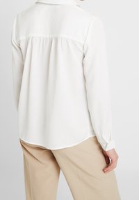 White long-sleeve shirt featuring a pleated back, soft texture, and a classic collar. Worn with light beige trousers.