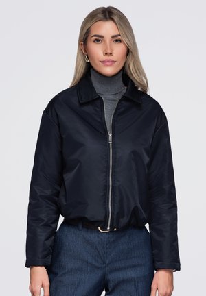 Femme aux longs cheveux blonds portant des boucles d'oreilles créoles en or, une veste zippée bleu foncé, un col roulé gris et un pantalon bleu, debout sur un fond blanc.