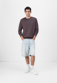 Mørk lilla sweatshirt med rund hals og en lille gul logo på brystet, parret med lyseblå denimshorts og hvide sneakers.