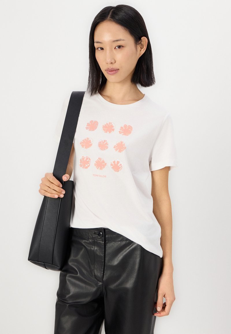 Femme avec des cheveux foncés raides portant un t-shirt blanc à imprimé floral, un pantalon en cuir noir et portant un sac à main noir sur fond blanc.