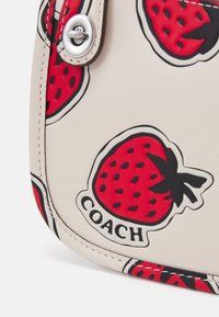 Coin de sac beige clair avec des motifs rouges de fraises et un texte noir « COACH » près d'une fermeture à loquet argentée.