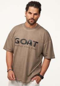 Braunes, übergroßes T-Shirt aus Baumwollmischung mit schwarzem "GOAT"-Druck und "GREATEST OF ALL TIME" darunter. Mit kurzen Ärmeln und rundem Halsausschnitt.